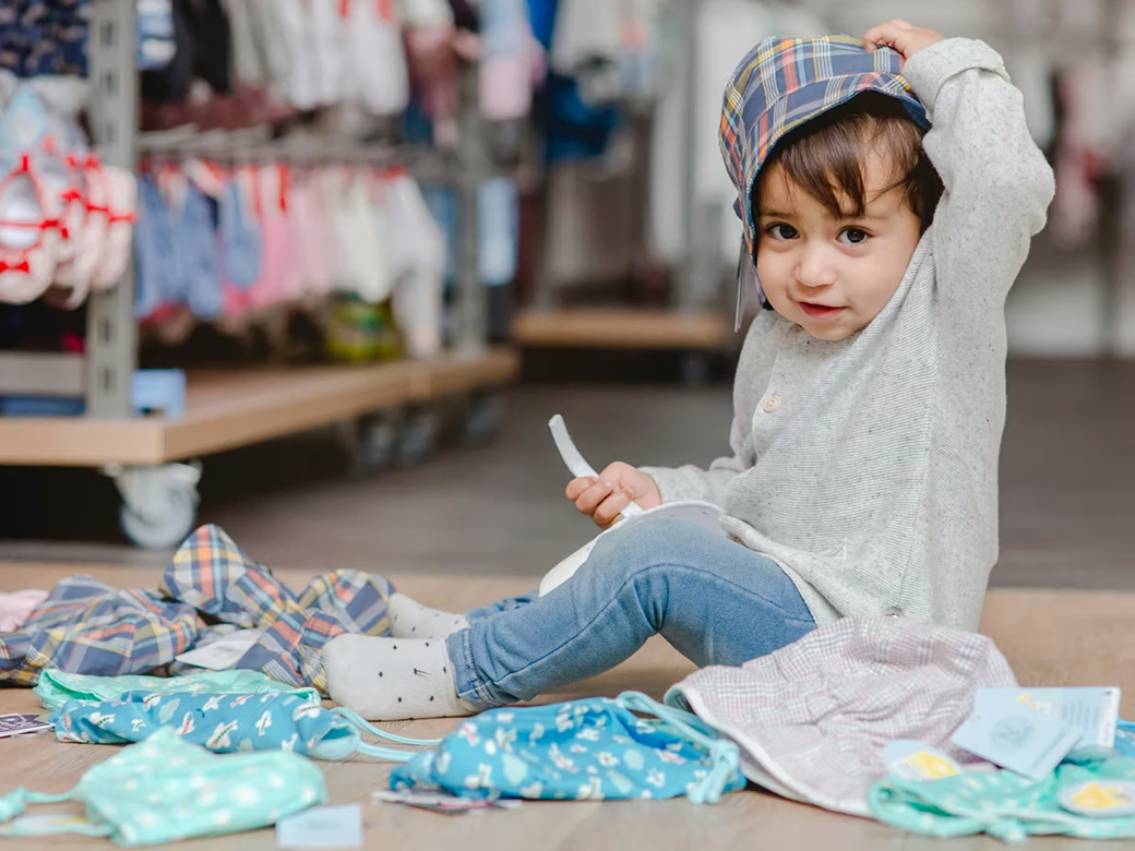 Kind sitzt auf dem Boden und hält eine Cap über seinen Kopf. Bekleidung liegt um das Kind herum.