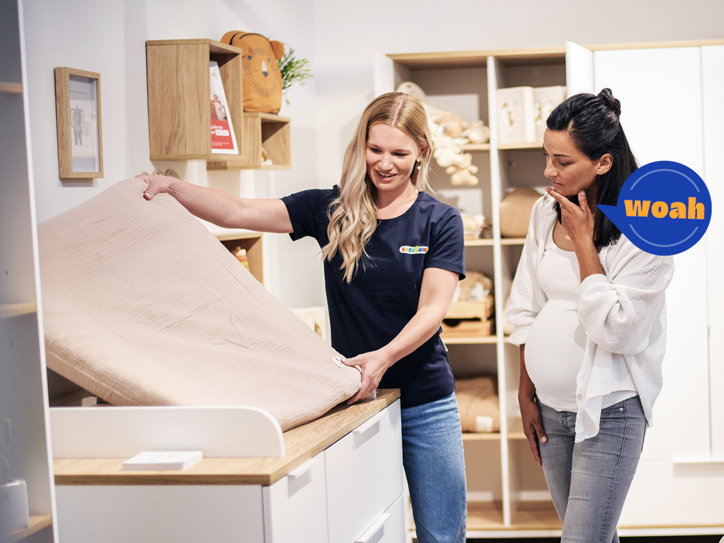 Eine Verkäuferin und eine Kundin schauen sich eine Wickelauflage an. Sprechblase bei Kundin mit Woah