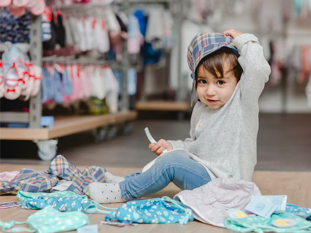 Kind sitzt auf dem Boden und hält eine Cap über seinen Kopf. Bekleidung liegt um das Kind herum.