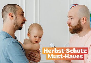 Herbst-Schutz Must-Haves: Vater mit Baby auf dem Arm. Ein anderer Mann reicht Medizin: Hausapotheke.
