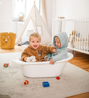 Zwei Kleinkinder sitzen in Bademänteln spielerisch in einer Wanne.