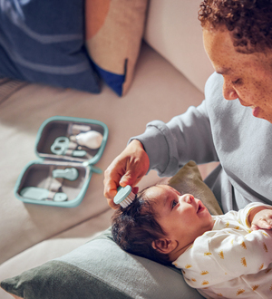 Eine Mutter kämmt vorsichtig die Haare eines Neugeborenen mit einer weichen Babybürste; daneben liegt ein Maniküre-Set.